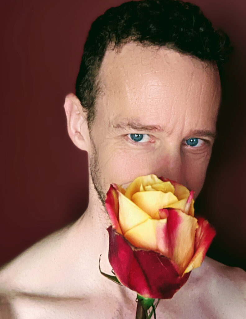 handsome white male, shirtless, holding a rose and coyly daring you to be joyful