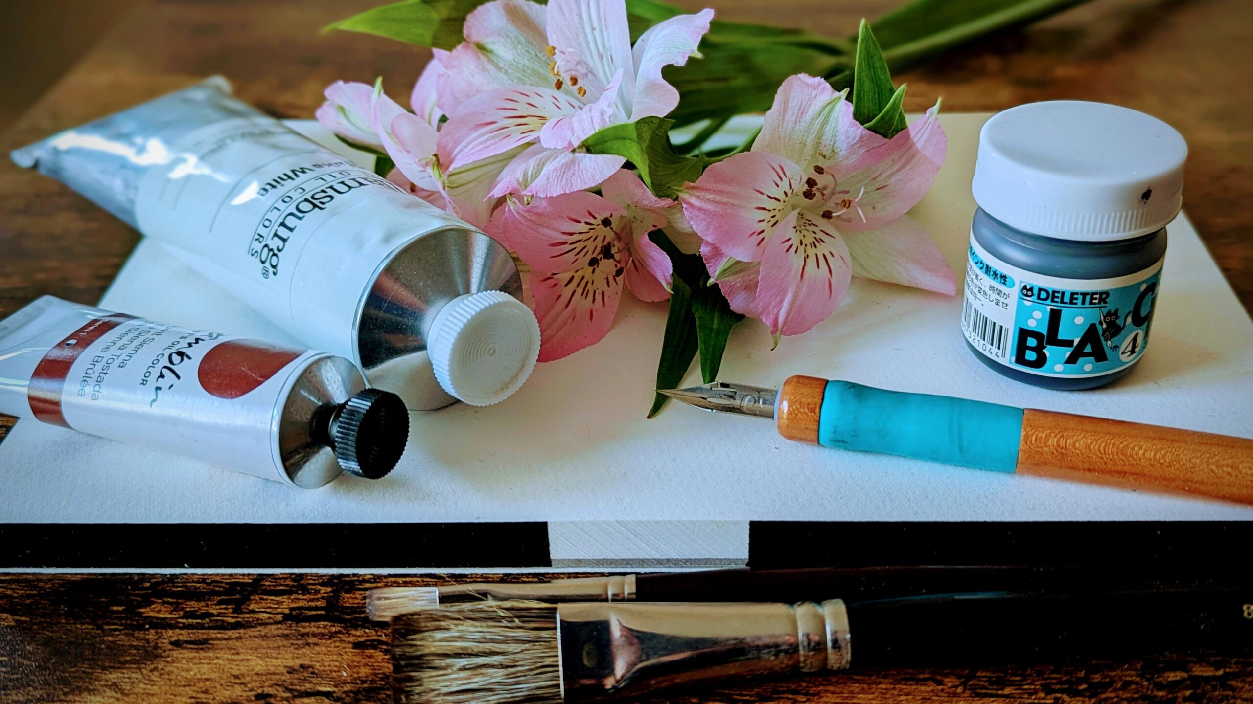 close up assemblage of painting and inking implements setting on a blank piece of white paper and decorated with a delicate pink bunch of flowers